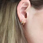 2 boucles d'oreilles en or blanc 18 carats en forme de créole sertie de diamants ainsi qu'une barrette en or rose sertie d'un grenat vert, ainsi qu'une autre en or rose perlé sertie d'un saphir rose, porté sur l'oreille d'un modèle.