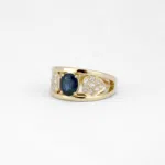 Bague en or jaune 18 carats composée de deux anneaux en forme de v comprenant un saphir bleu au centre de ces anneaux et de deux pétales de fleurs serties de diamants autour de ce saphir bleu.
