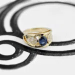Bague en or jaune 18 carats composée de deux anneaux en forme de v comprenant un saphir bleu au centre de ces anneaux et de deux pétales de fleurs serties de diamants autour de ce saphir bleu.