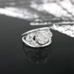 Bague en or blanc 18 carats composée de deux anneaux en forme de v comprenant un diamant au centre de ces anneaux et de deux fleurs en forme de diamants autour de ce saphir bleu.