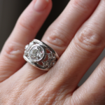 Bague en or blanc 18 carats composée de deux anneaux en forme de v comprenant un diamant au centre de ces anneaux et de deux fleurs en forme de diamants autour de ce saphir bleu. Porté sur une main.