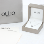 Ecrin de bijou contenant un pendentif en or blanc 18 carats serti d'un diamant entouré de diamants, ainsi qu'une chaîne en or blanc 18 carats. Un sac de shopping gris avec le logo Olijo et le certificat d'authenticité.
